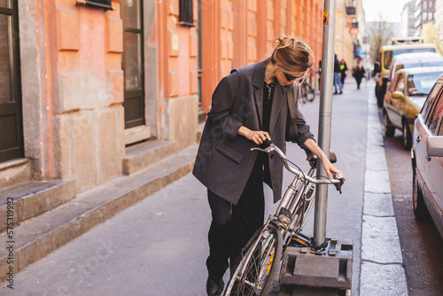 Attractive business woman come to sits on bike. Girl wear grey jacket or blazer, black pants, eyeglasses. Modern business woman ride to work on vehicle. Way to office. Riding bicycle to work. © zvkate