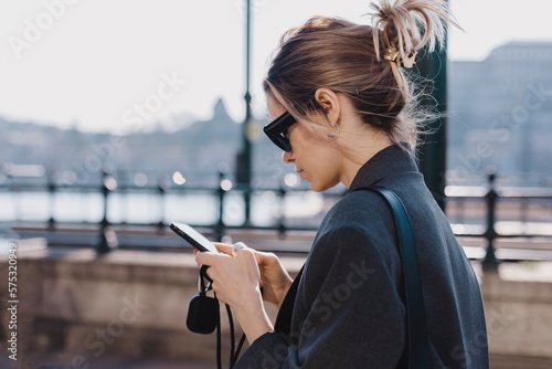 Photo of young woman dreaming, reading messages on a smartphone while going on embankment Fototapet