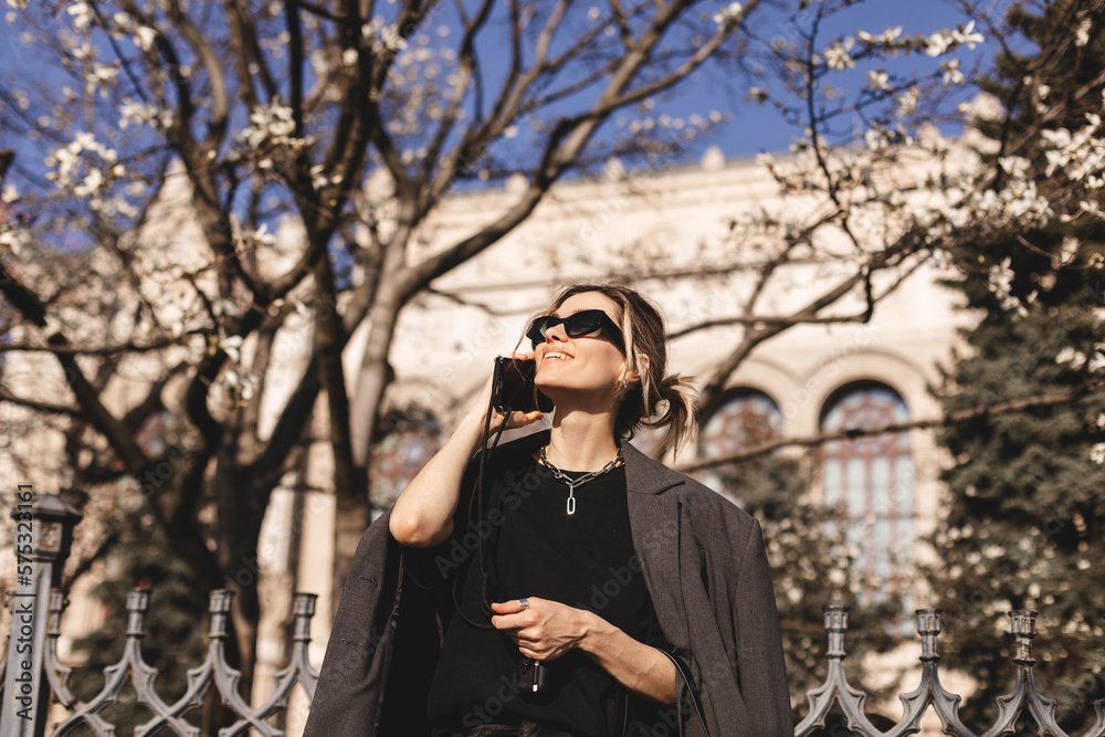 Beautiful young woman wearing suit, walking outdoors, talking on mobile phone. Beautiful Woman talking on phone. Portrait of stylish smiling business woman calling on mobile phone under blooming tree. Beautiful young woman wearing suit, walking outdoors, talking on mobile phone. Beautiful Woman talking on phone. Portrait of stylish smiling business woman calling on mobile phone under blooming tree.