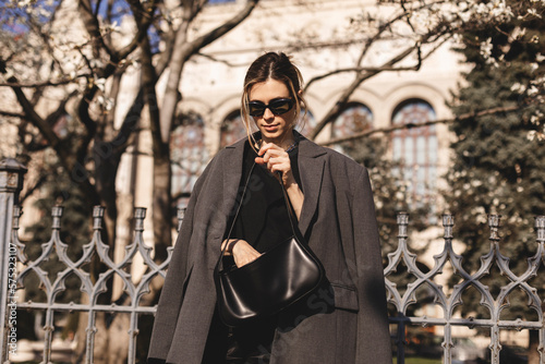 Elegant young woman looking in her black leather bag her phone or purse. Business style woman wear grey blazer, black eyeglasses and bag on the street. Street style, fashion outfit.