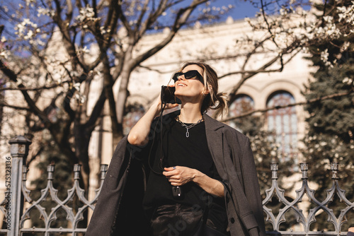 Beautiful young woman wearing suit, walking outdoors, talking on mobile phone. Beautiful Woman talking on phone. Portrait of stylish smiling business woman calling on mobile phone under blooming tree.