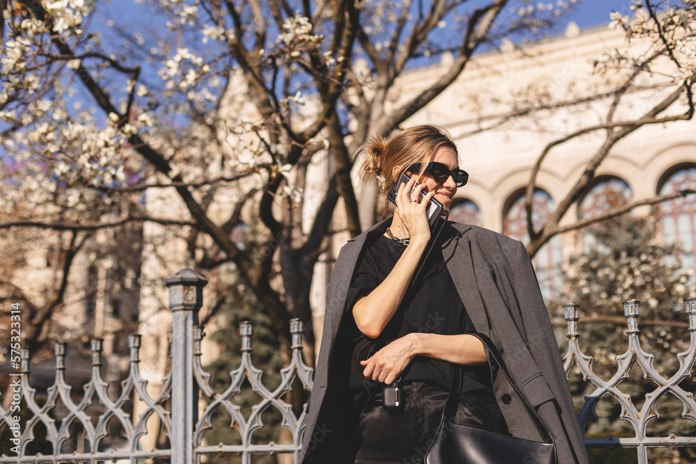 Beautiful young woman wearing suit, walking outdoors, talking on mobile phone. Beautiful Woman talking on phone. Portrait of stylish smiling business woman calling on mobile phone under blooming tree. Beautiful young woman wearing suit, walking outdoors, talking on mobile phone. Beautiful Woman talking on phone. Portrait of stylish smiling business woman calling on mobile phone under blooming tree.