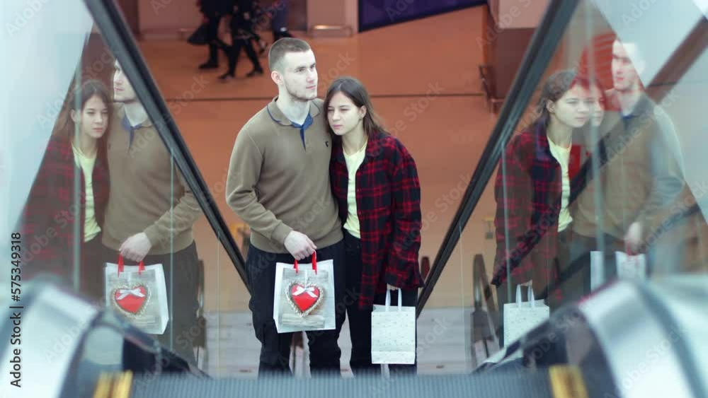 Happy couple in love in shopping mall is climbing escalator with paper bags and friendly communicate. Young people are walking and shopping at the mall. Love, holiday sales, retail, consumerism. Happy couple in love in shopping mall is climbing escalator with paper bags and friendly communicate. Young people are walking and shopping at the mall. Love, holiday sales, retail, consumerism.
