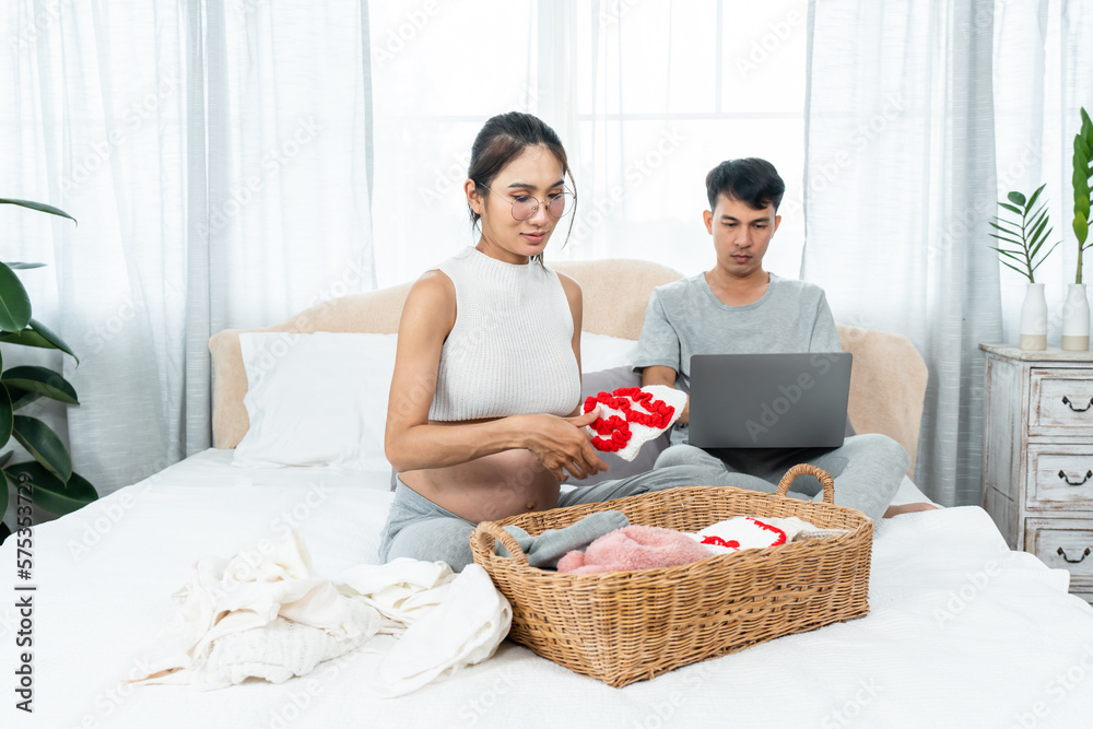 Asian teen mom is pregnant, pregnant, maternity, sitting and preparing clothes for upcoming baby, baby clothes, in mother's hand, on bed in bedroom, husband sitting smiling happily in back