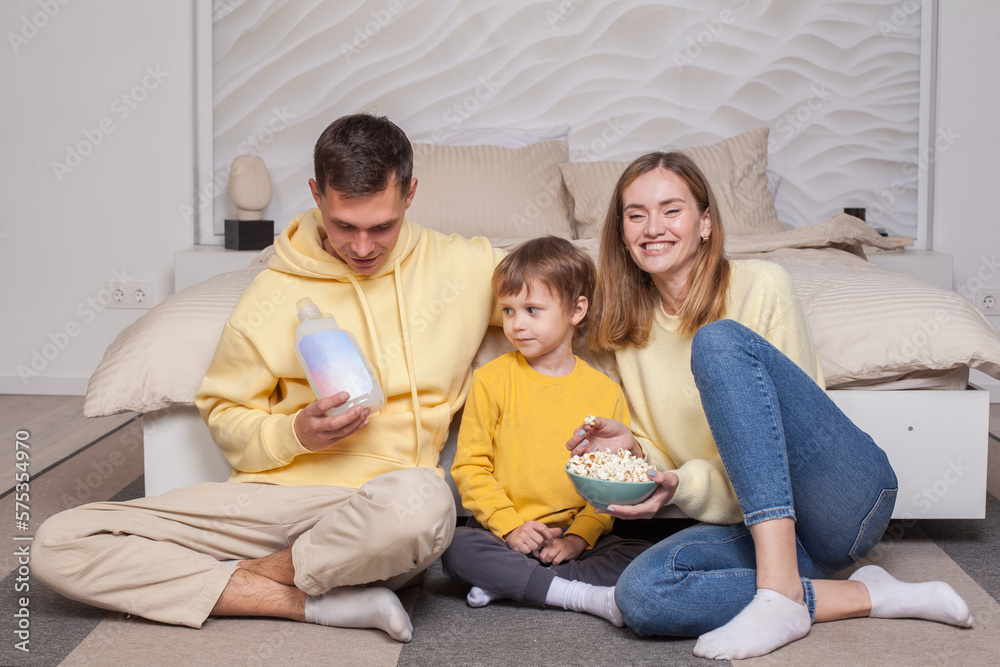 Cute adorable smiling parents with child son sitting by the bed and eating popcorn, dad holding bottle, advertising Cute adorable smiling parents with child son sitting by the bed and eating popcorn, dad holding bottle, advertising