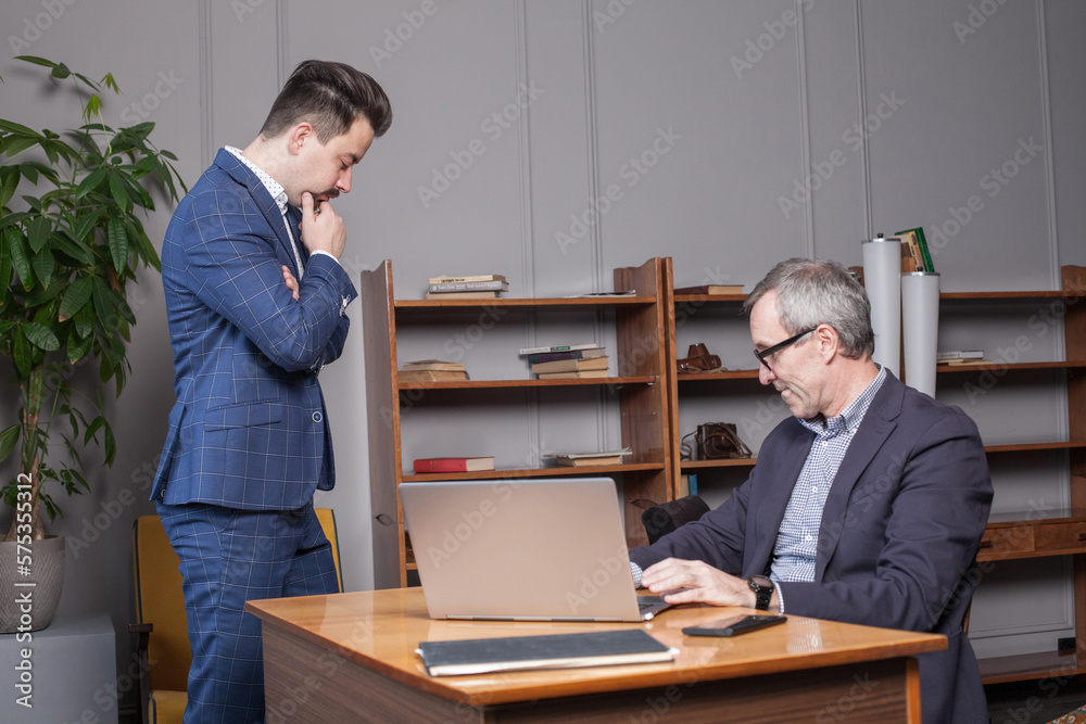 Mature serious businessman in dark blue suit with his partner are standing by laptop in office and thinking about projects Mature serious businessman in dark blue suit with his partner are standing by laptop in office and thinking about projects