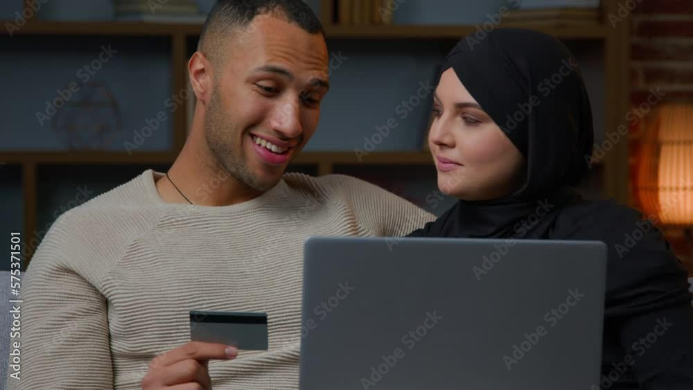 Ethnic spouses couple use credit card with laptop African american man muslim woman in hijab paying online electronic bank application multiracial family shoppers spending money on internet e-commerce Ethnic spouses couple use credit card with laptop African american man muslim woman in hijab paying online electronic bank application multiracial family shoppers spending money on internet e-commerce