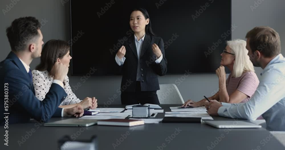 Asian business coach makes presentation, share information to team, looks confident leads formal meeting with company investors or clients sit at desk gathered in boardroom. Corporate training event