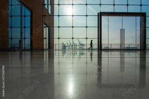 Blick durch ein großes modernes Glasfenster, rechts eine große Glastür, links im Bild Teil des rotbraunen Gebäudes, im Hintergrund die Silhouetten von Hafenkränen und einer Joggerin zu sehen, 