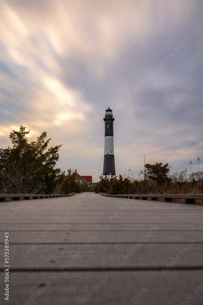Wooden boardwalk leading to a tall stone lighthouse with moody streaky clouds in the sky. Fire Island, Long Island New York
