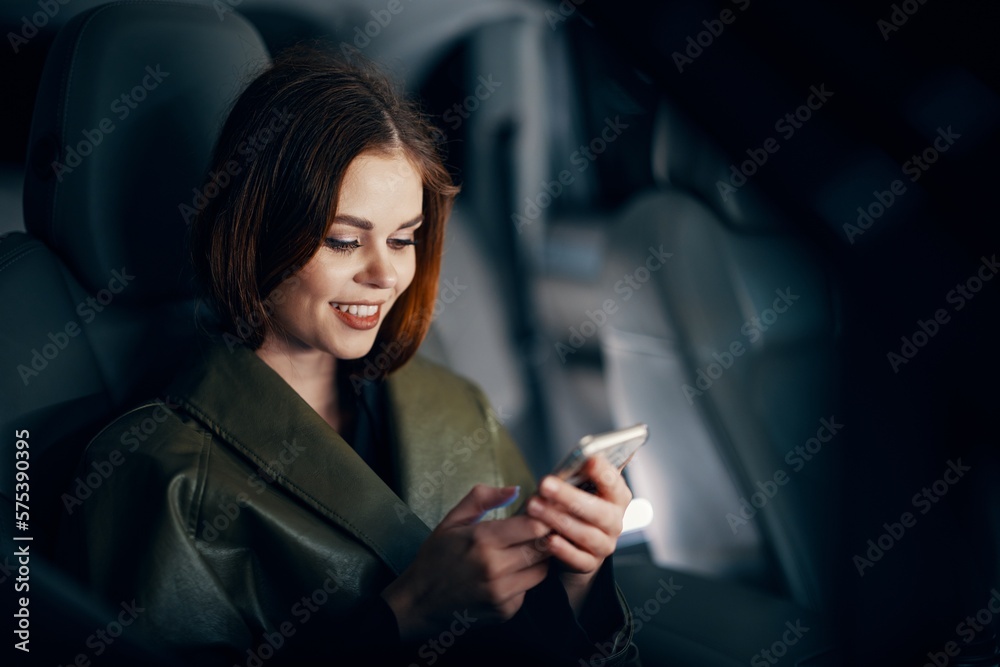 a close horizontal portrait of a stylish, luxurious woman in a leather coat sitting in a black car at night in the passenger seat, happily looking at her phone during the trip a close horizontal portrait of a stylish, luxurious woman in a leather coat sitting in a black car at night in the passenger seat, happily looking at her phone during the trip