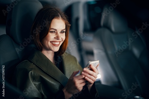 horizontal portrait of a stylish, luxurious woman in a green leather coat, sitting in a black car at night on the passenger seat, smiling happily, holding her phone during the trip © SHOTPRIME STUDIO