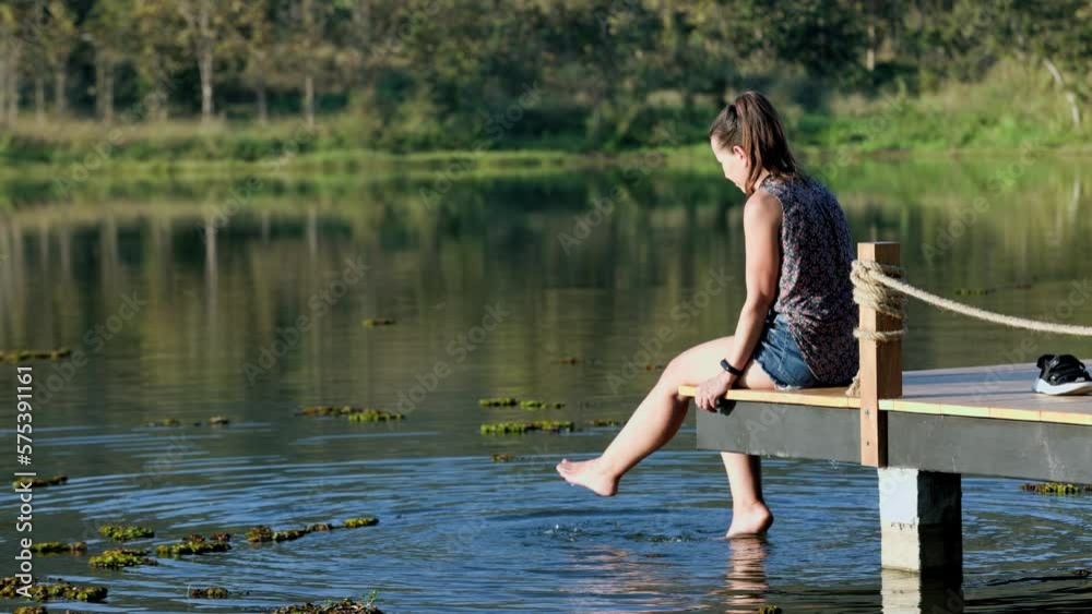 A woman in a blouse sits on a wooden pier by the lake in summer or autumn with bright trees and reflections in the water A woman in a blouse sits on a wooden pier by the lake in summer or autumn with bright trees and reflections in the water