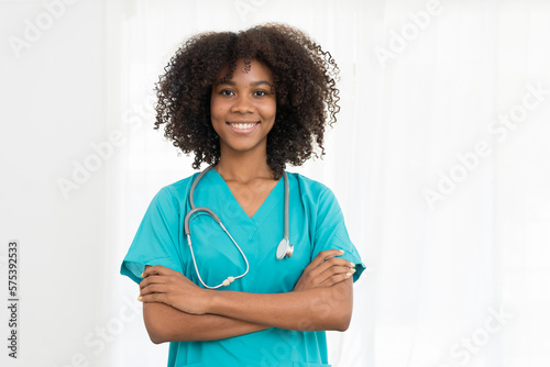 Portrait of smiling young female doctor or young nurse wearing blue scrubs uniform and stethoscope and standing with arms crossed while looking at camera isolated on white background
