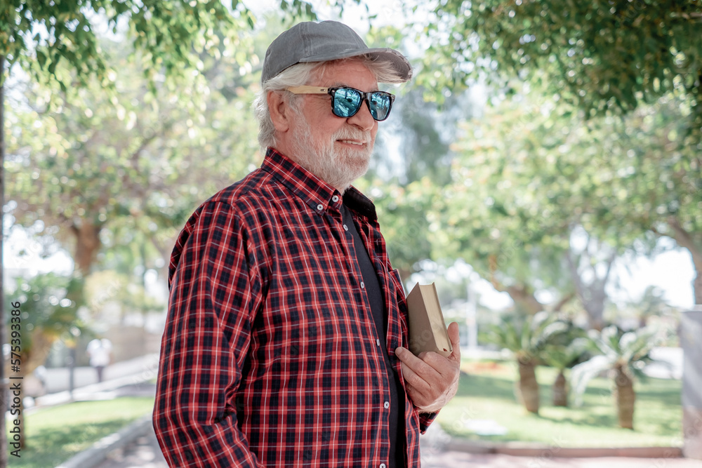 Handsome senior bearded man walking in urban park with a book under his arm. Smiling pensioner enjoying free time and retirement