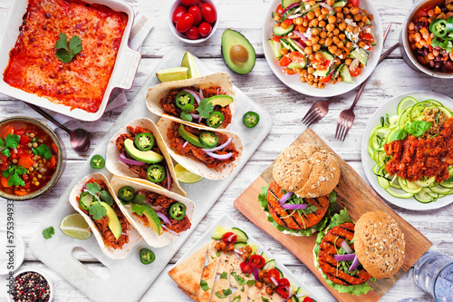 Healthy plant based vegetarian meal table scene. Above view on a white wood background. Jackfruit tacos, zucchini lasagna, walnut bolognese zoodles, chickpea burgers, hummus, soups, salad.