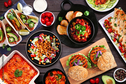 Healthy plant based vegetarian meal table scene. Overhead view on a dark wood background. Jackfruit tacos, zucchini lasagna, walnut bolognese zoodles, chickpea burgers, hummus, soups, salad.