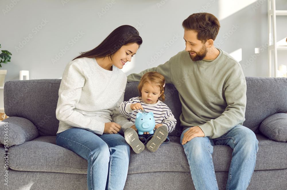 Loving, happy, young family with their little daughter are putting coins together in piggy bank, sitting at home on the sofa in the living room. Earnings, saving, currency and finance concept. Loving, happy, young family with their little daughter are putting coins together in piggy bank, sitting at home on the sofa in the living room. Earnings, saving, currency and finance concept.