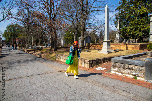 an African American woman wearing a yellow dress, a colorful orange head scarf, a denim jacket with long sisterlocks surrounded by lush green trees at the Oakland Cemetery in Atlanta Georgia USA photo