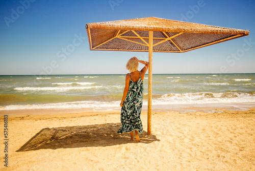 The girl stands facing the ocean near the beach umbrella.