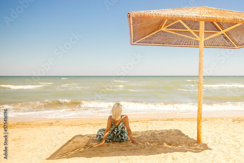 The girl sits facing the ocean, hiding from the sun under a beach umbrella.