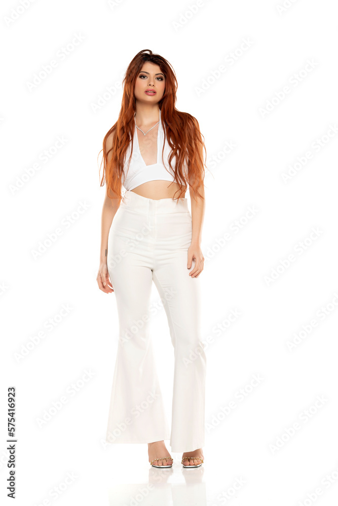 Fashion style studio portrait of beautiful young woman. White wide leg pants and white sleevles shirt. Model standing and posing against white background Fashion style studio portrait of beautiful young woman. White wide leg pants and white sleevles shirt. Model standing and posing against white background
