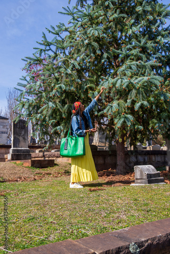 an African American woman wearing a yellow dress, a colorful orange head scarf, a denim jacket with long sisterlocks with lush green trees, headstones and graves at the Oakland Cemetery in Atlanta photo