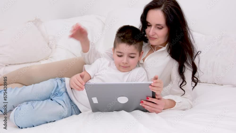 mom and son are lying comfortably on the bed relaxing in a beautiful white hotel playing a game on a tablet calling home have gone to another country white clothes black hair love caring motherhood