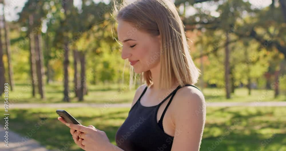 A blonde woman in black sportswear stands in a park with a phone in her hands, smiles and looks at the smartphone screen. A blonde woman in black sportswear stands in a park with a phone in her hands, smiles and looks at the smartphone screen.