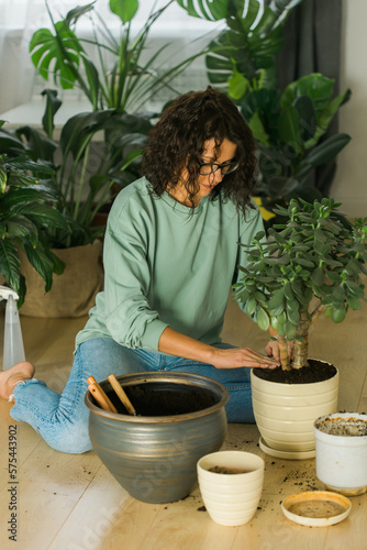 Smiling young woman and pot with plant happy work in indoor garden or cozy home office with different houseplants. Happy millennial female gardener or florist take care of domestic flower
