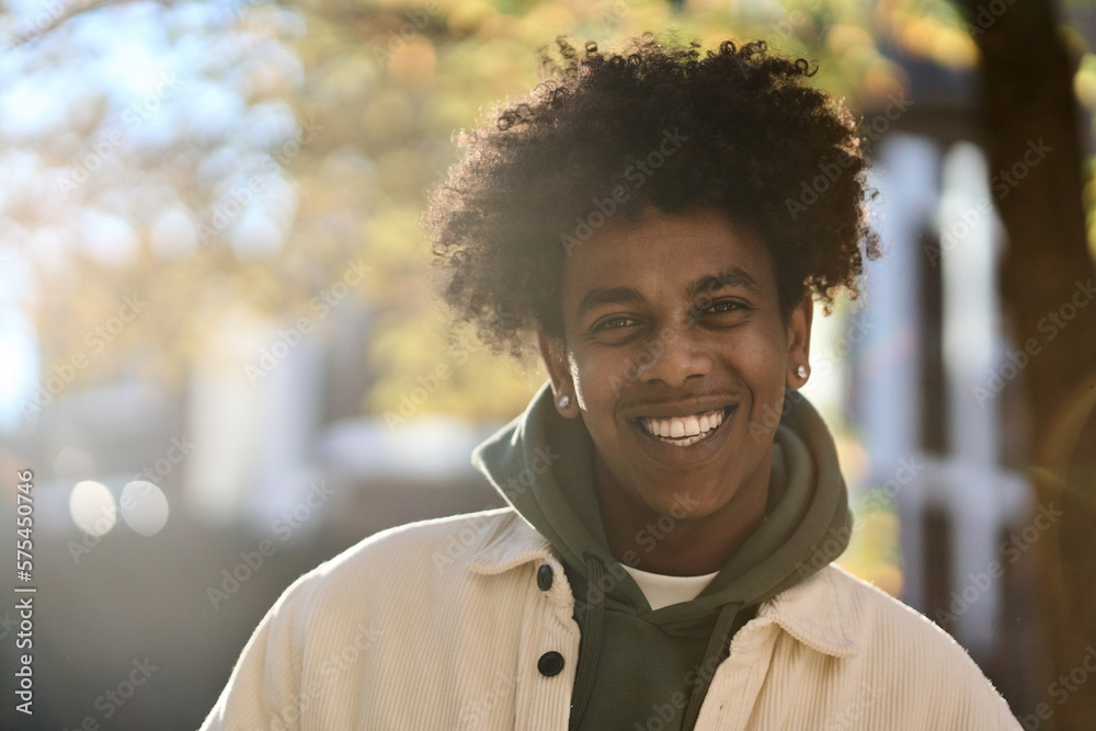 Smiling positive young African American guy model standing at big city sunny street. Stylish ethnic hipster gen z teenager feeling happy looking at camera outdoors in park, Close up portrait. - obrazy, fototapety, plakaty 