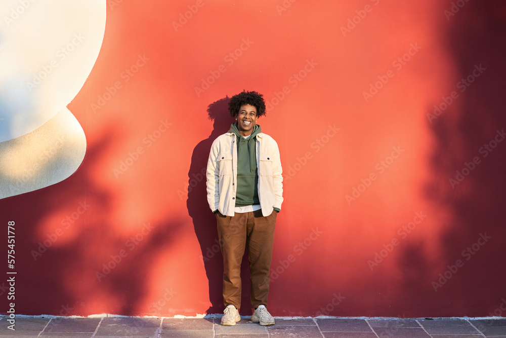 Happy African American teen guy having fun laughing on red wall lit with sunlight. Smiling cool ethnic gen z teenager model standing looking at camera, authentic full length shot outdoors. - obrazy, fototapety, plakaty 