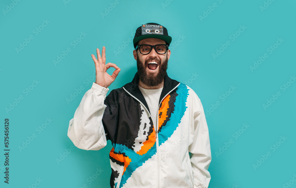 Fashionable bright man with a beard in a vintage sports jacket, cap and glasses with an audio cassette in retro style isolated on a blue studio background. Old school Fashionable bright man with a beard in a vintage sports jacket, cap and glasses with an audio cassette in retro style isolated on a blue studio background. Old school
