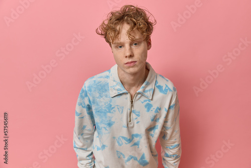 Portrait of curly red haired stylish smart teenager with a perfect skin wearing colorful clothing shirt looking at camera posing stands leaning over against pink backdrop copy space in a studio shot.