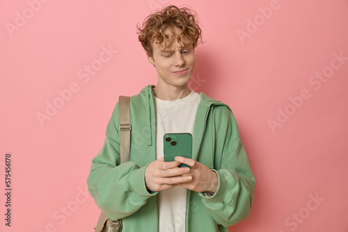 Fotografi Portrait of puzzled amazed curly haired male standing in a studio shot, wearing green zipped hoodie and holding a smartphone while reading embarrassing text messages isolated over pink background