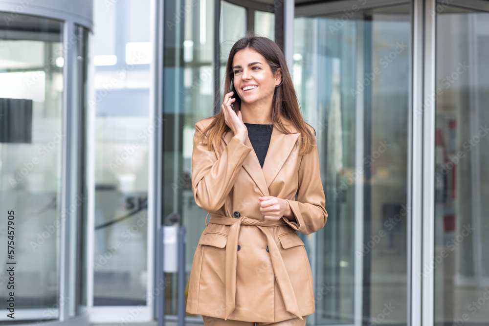 Fototapeta premium Smiling successful young businesswoman leader entrepreneur, CEO or professional manager in a stylish business attire talking using mobile phone leaving the business center