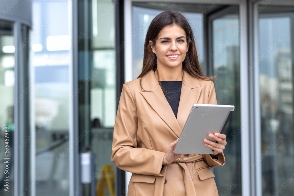 Smiling successful young business woman in stylish business attire holding digital tablet computer against the backdrop of the business center looks at the camera and smiles friendly, success concept Smiling successful young business woman in stylish business attire holding digital tablet computer against the backdrop of the business center looks at the camera and smiles friendly, success concept
