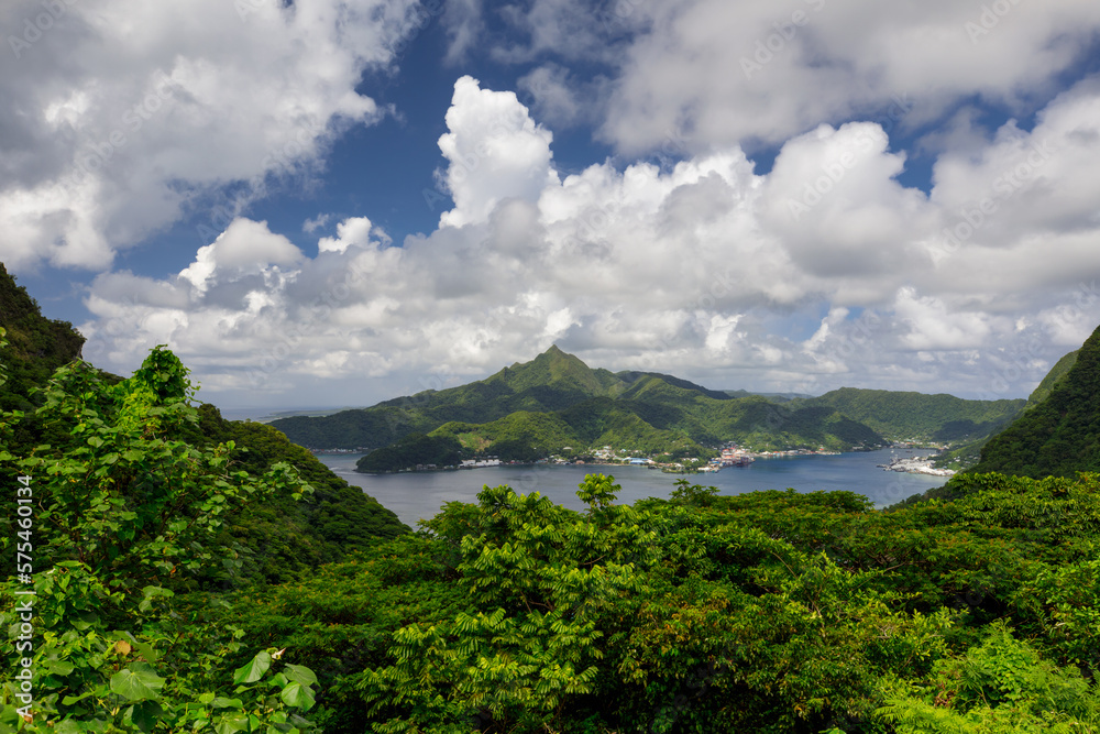 A scenic view of Pago Pago Harbor and Mt. Matafau with the lush green foliage framing the scene and white fluffy clouds in the beautiful blue sky.