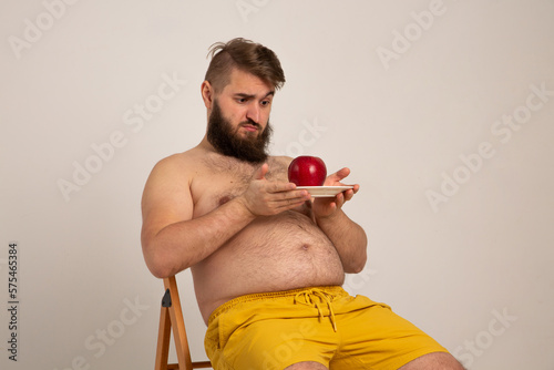 Young bearded man sits on wooden chair and looks incredulously at an apple. A man with a beard and overweight problem chooses a fruit diet. A fat man with big belly wants to switch to a healthy diet photo