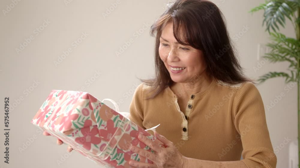 Smiling attractive mature woman in brown holding colorfully wrapped present is happy and surprised shaking box and making delightful expressions at gift. Beautiful middle age asian female enjoys. Smiling attractive mature woman in brown holding colorfully wrapped present is happy and surprised shaking box and making delightful expressions at gift. Beautiful middle age asian female enjoys.