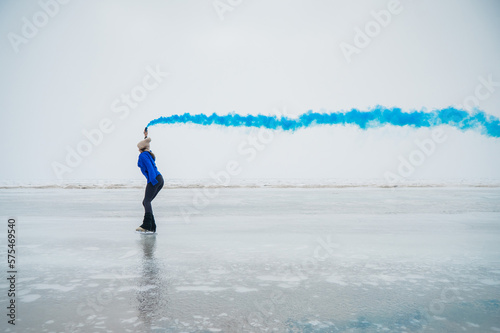 A caucasian woman is skating on a frozen lake holding colored blue smoke in her hands. © Михаил Решетников