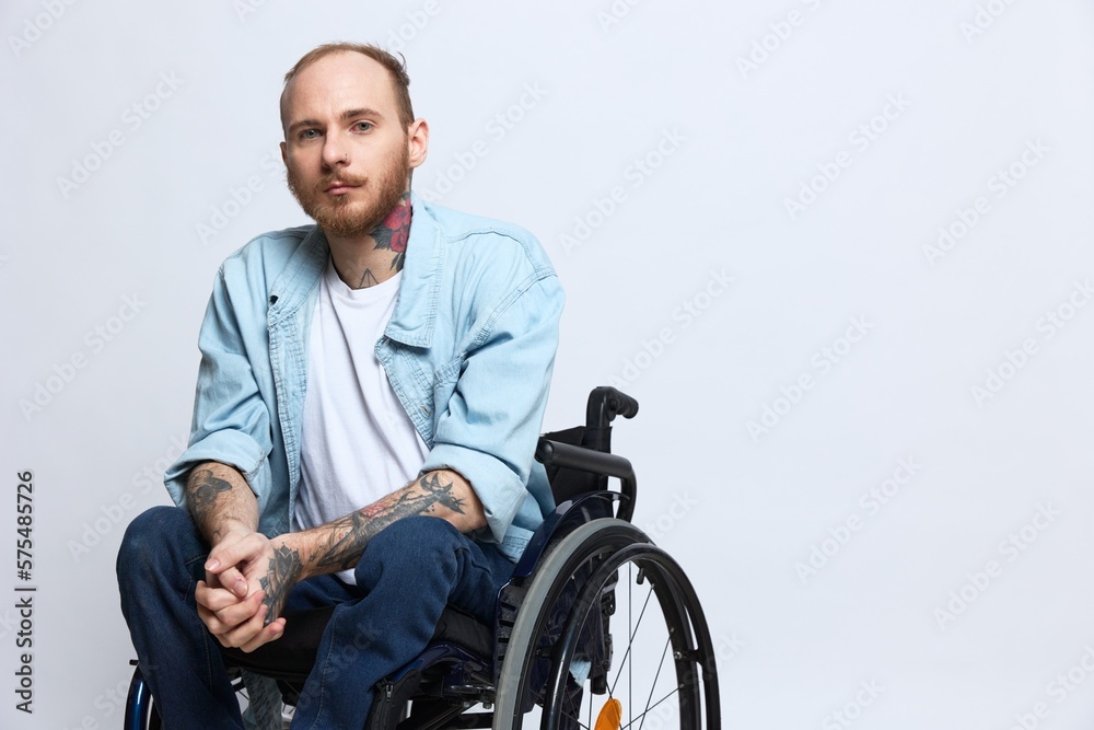 A man in a wheelchair looks thoughtfully at the camera, copy space, with tattoos on his arms sits on a gray studio background, the concept of health a person with disabilities, a real person A man in a wheelchair looks thoughtfully at the camera, copy space, with tattoos on his arms sits on a gray studio background, the concept of health a person with disabilities, a real person