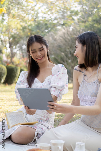 Two young Asian women are using tablet, taking selfies with tablet or watching videos on a digital tablet together while enjoying a picnic in the park