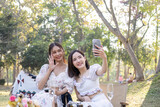 Two beautiful young Asian women picnic in the green park together, taking selfies with their phone they are having picnic,they talk happily