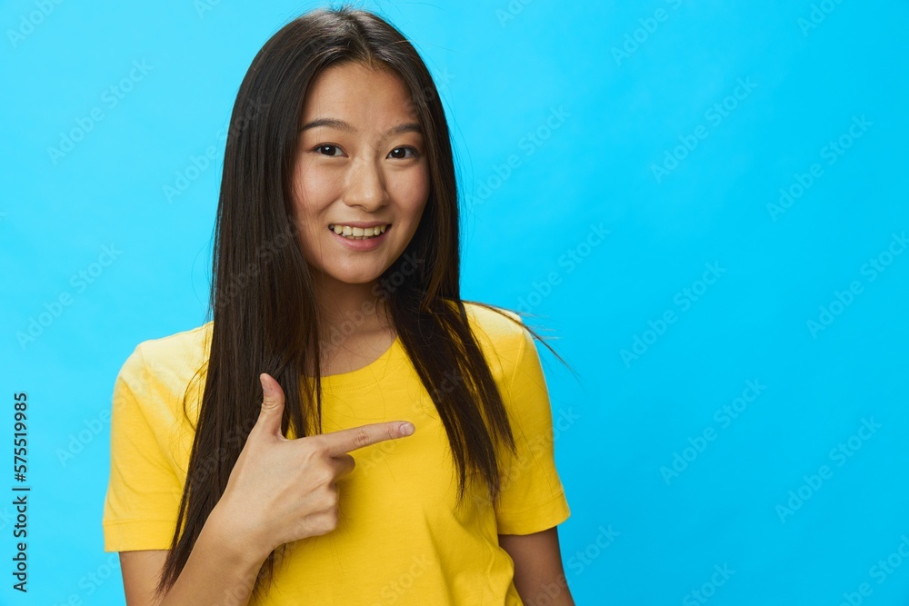 Asian woman points to a loose background in a yellow T-shirt and blue jeans on a blue background