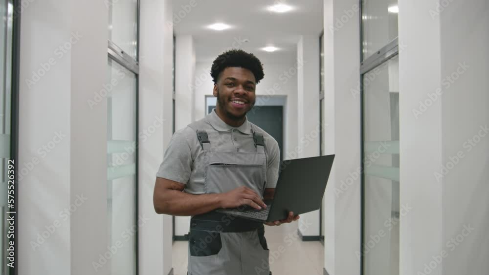 African CCTV cameras installer in uniform stands in corridor, smiles and looks at camera. He sets up security cameras in office using software on laptop. Concept of surveillance system. Dolly zoom. African CCTV cameras installer in uniform stands in corridor, smiles and looks at camera. He sets up security cameras in office using software on laptop. Concept of surveillance system. Dolly zoom.