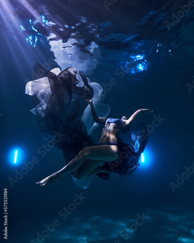 Fototapeta Naklejka Na Ścianę i Meble -  a young beautiful girl is swimming in a pool under water fashion fabric on a dark blue background like in the sea