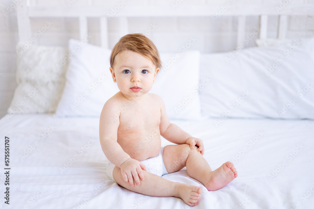 a joyful baby girl of six months on a bed in diapers smiles, a small child on a cotton bed at home, the concept of care and hygiene a joyful baby girl of six months on a bed in diapers smiles, a small child on a cotton bed at home, the concept of care and hygiene