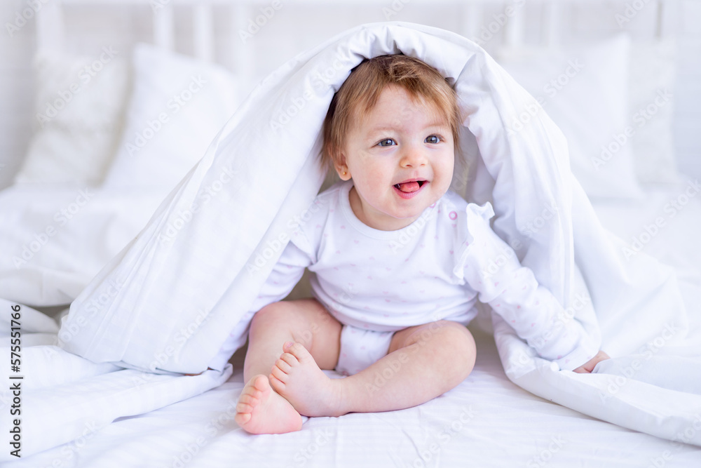funny happy baby girl looks out from under the blanket on the bed in white clothes and smiles, a small child on a cotton bed at home woke up in the morning after sleeping funny happy baby girl looks out from under the blanket on the bed in white clothes and smiles, a small child on a cotton bed at home woke up in the morning after sleeping