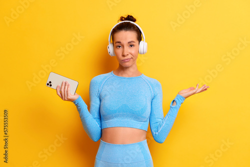 Indoor shot of disappointed upset fit woman in trendy sportswear holds modern smartphone keeps palm aside has puzzled expression can not browse music app poses with white headphones isolated over photo